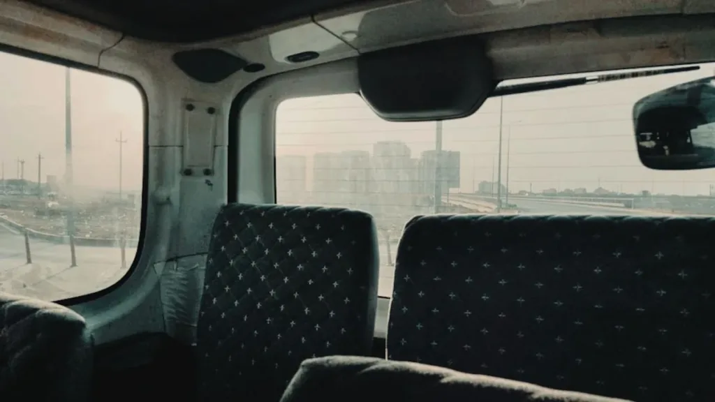 Empty rear passenger seats inside a van, rows of dark patterned upholstery with windows and a hazy city view beyond