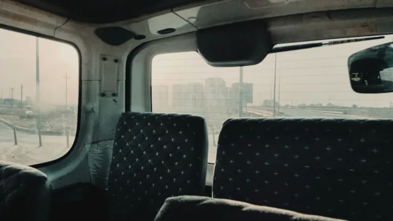 Empty rear passenger seats inside a van, rows of dark patterned upholstery with windows and a hazy city view beyond