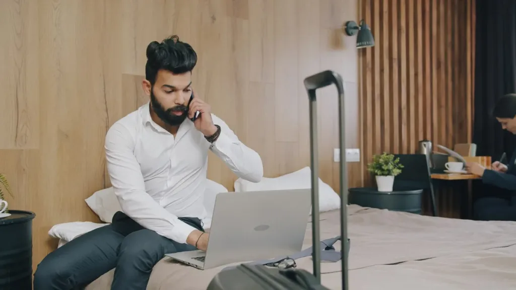 Business traveller on a phone call with a laptop on a hotel bed, a wheeled suitcase standing beside him