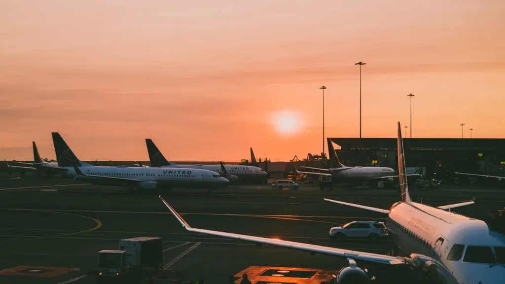Passenger jets parked on an airport apron at sunset, with service vehicles and floodlight masts under an orange sky