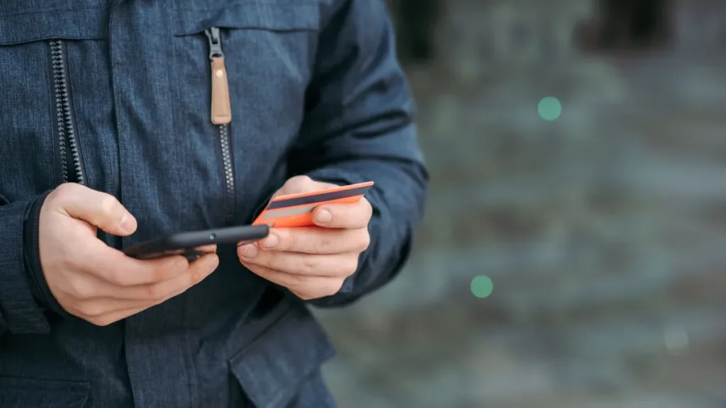 Hands of a person in a dark jacket holding a smartphone and a bank card, about to enter card details for an online payment