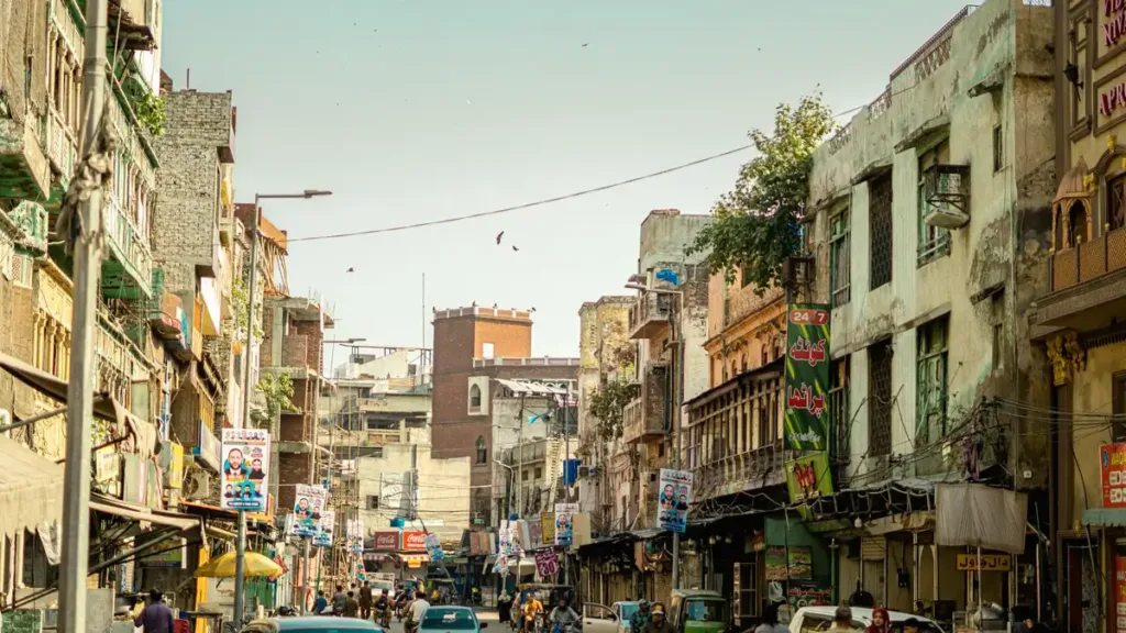 Busy Lahore street lined with older multi-storey buildings, shops and small cars moving and parked along the kerb