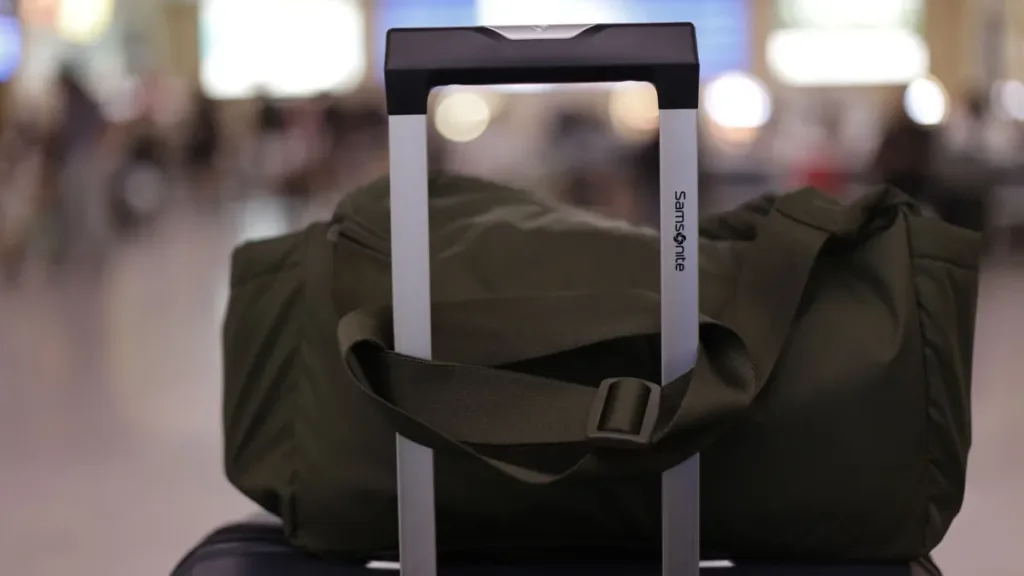 Dark green duffel bag resting on a wheeled suitcase with its handle raised, in a softly blurred airport terminal