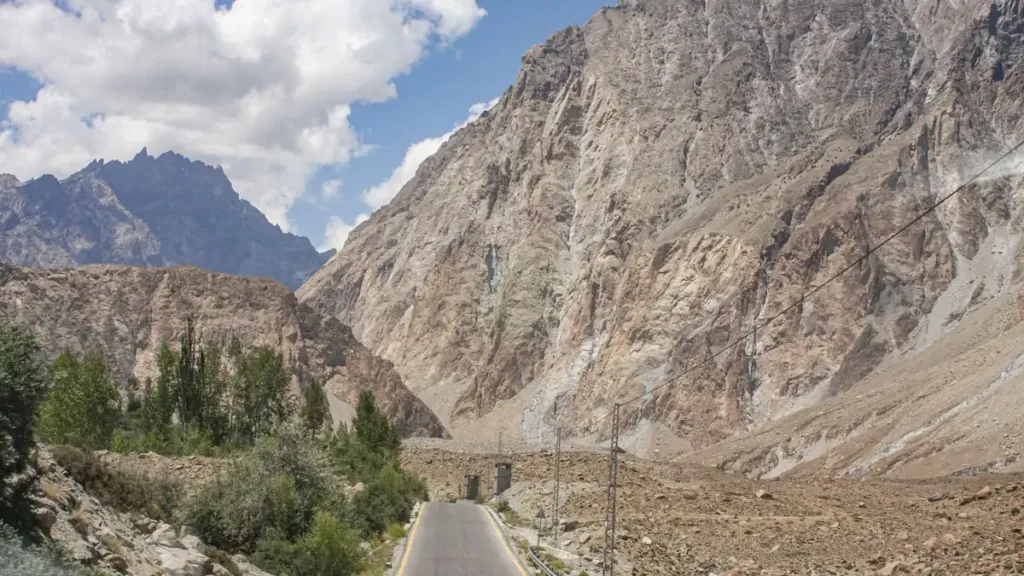 Empty paved mountain road winding between steep rocky slopes and green trees under a blue sky with clouds