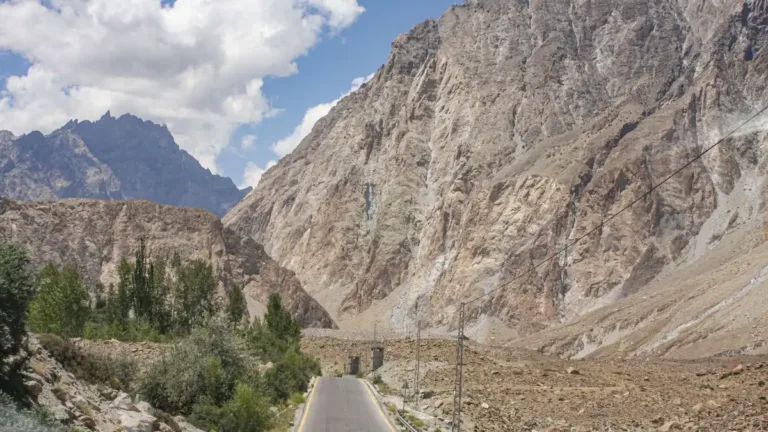 Empty paved mountain road winding between steep rocky slopes and green trees under a blue sky with clouds