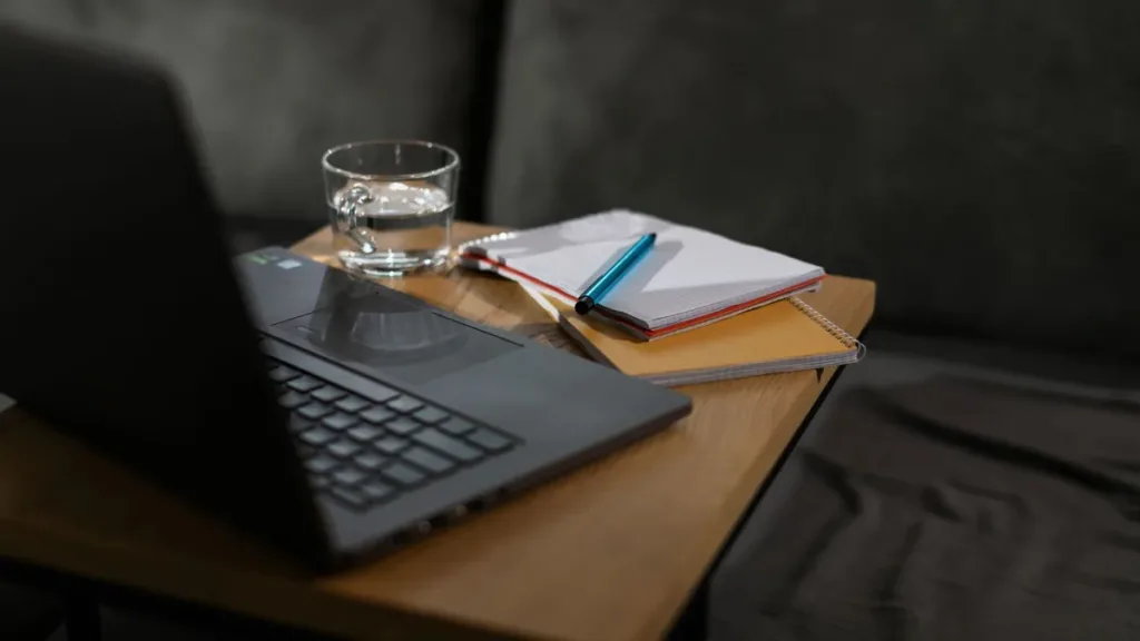 Open laptop beside notebooks, a pen and a glass of water on a small wooden side table in a dim room