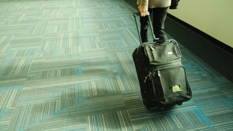 Traveller in dark trousers pulling a black wheeled carry-on case along a carpeted airport corridor