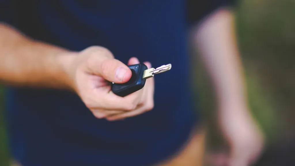 A hand holding out a black car key fob, face out of frame, against a softly blurred outdoor background