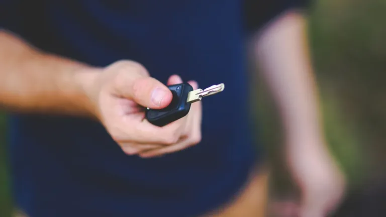 A hand holding out a black car key fob, face out of frame, against a softly blurred outdoor background
