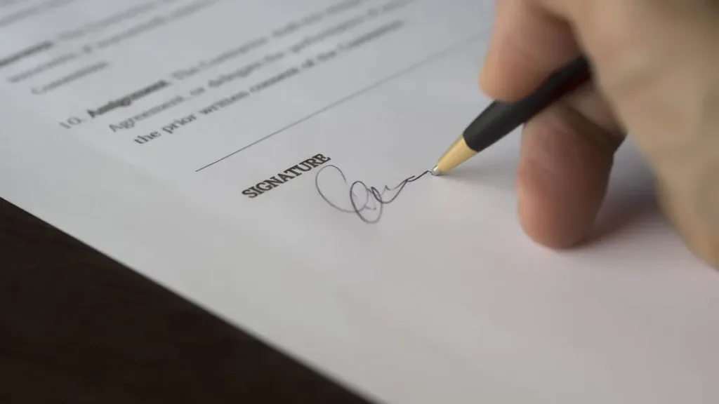 Close-up of a hand signing the signature line of a printed agreement with a pencil on a desk