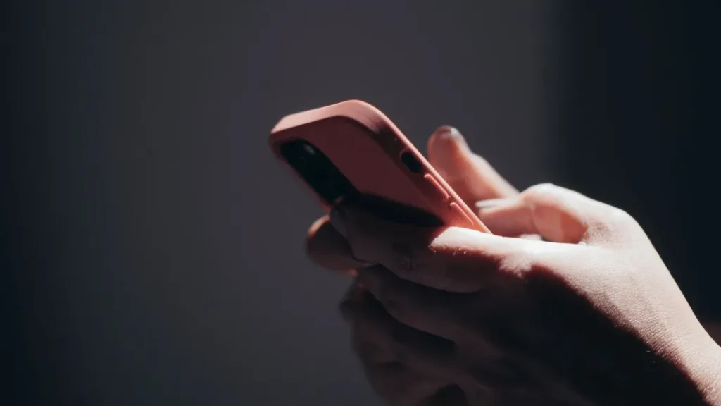 Close-up of two hands holding a coral-cased smartphone against a dark background, lit by a narrow beam of light