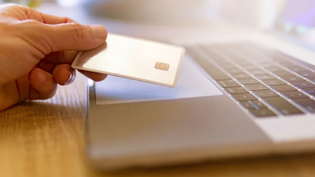 Hand holding a plain payment card beside an open laptop keyboard on a wooden desk, ready to pay online