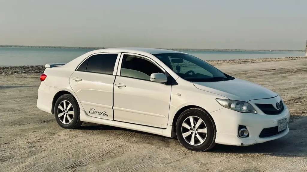 White Toyota Corolla saloon parked on a sandy beach with calm sea and a hazy sky behind it