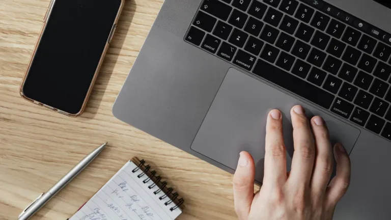 Overhead view of a hand on a laptop trackpad beside a smartphone, pen and spiral notepad on a wooden desk