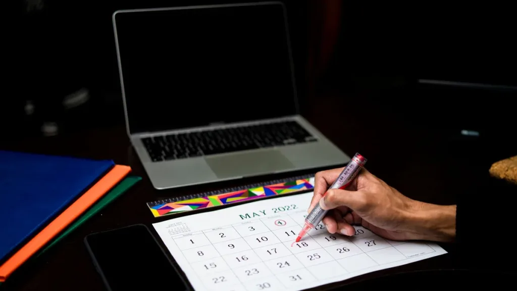 Hand marking dates on a paper desk calendar with a pink pen, next to an open laptop and a smartphone on a dark desk