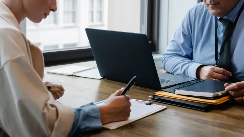 Two colleagues at an office desk with a laptop, one signing a paper form as a folder and tablet sit beside them