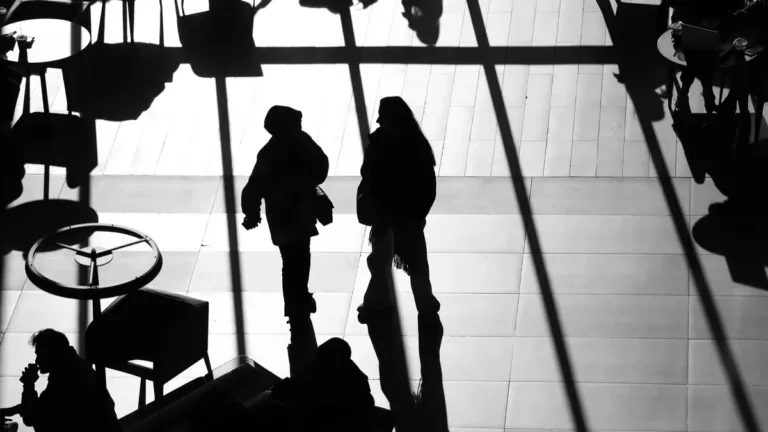 Black and white silhouettes of two travellers walking through a sunlit airport terminal past cafe tables and chairs