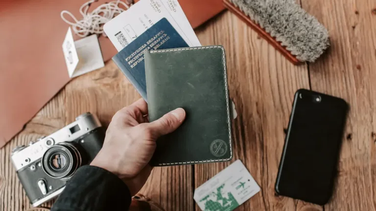 Hand holding a plain green leather passport wallet beside boarding passes, a phone and a film camera on a wooden table