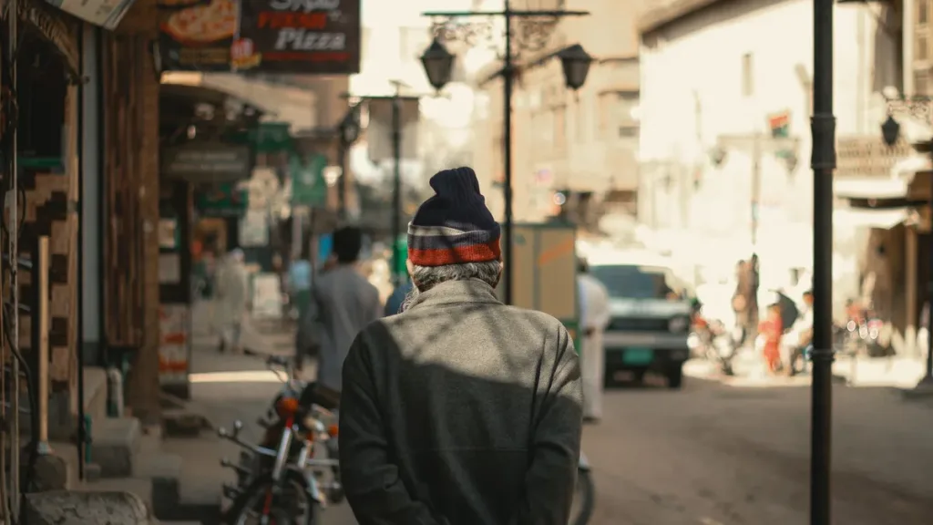 Man in a knit cap seen from behind on a Peshawar street lined with shops, signboards and parked motorbikes
