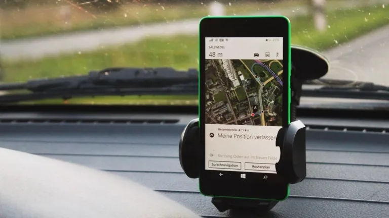 Smartphone in a dashboard cradle showing a satellite map with a location pin, seen through a car windscreen