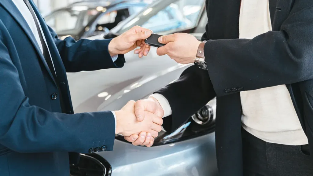 Two people in suits shake hands while one passes a car key to the other beside a silver car indoors