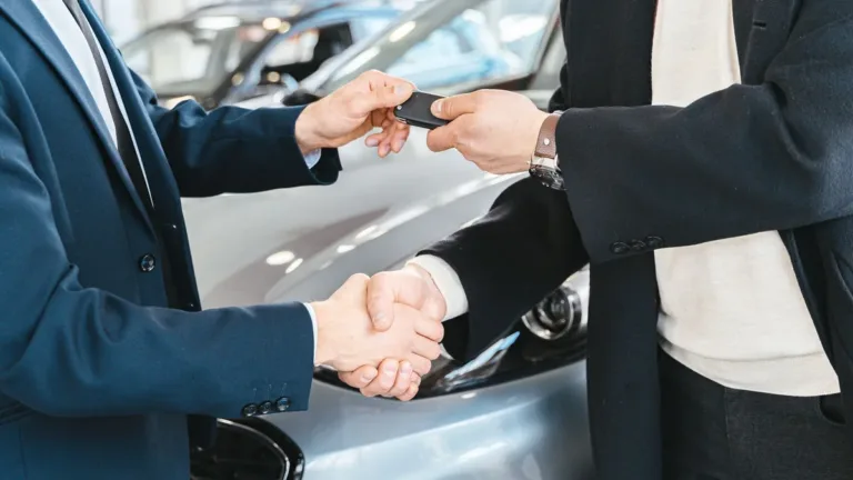Two people in suits shake hands while one passes a car key to the other beside a silver car indoors