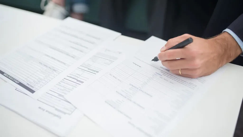 Close-up of a hand holding a pen over printed forms on a white desk, reviewing paperwork before signing