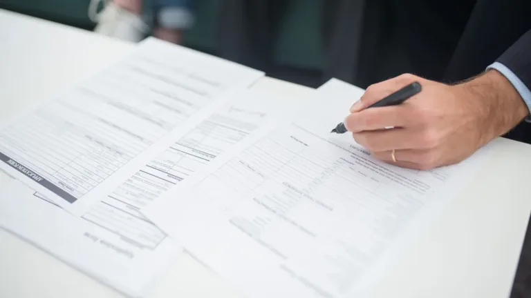 Close-up of a hand holding a pen over printed forms on a white desk, reviewing paperwork before signing