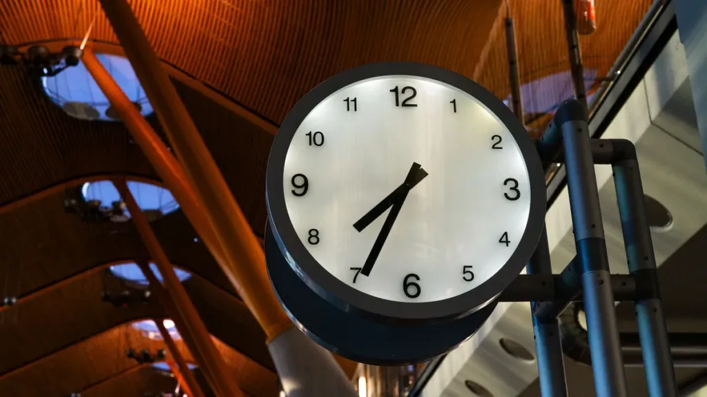 Large round analogue clock showing just after one o'clock under a curved wooden airport terminal ceiling