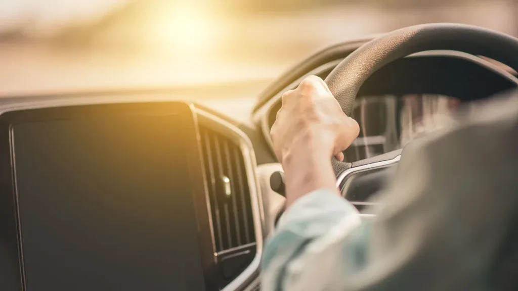 Driver's hand on the steering wheel of a right-hand-drive car, centre touchscreen to the left, low sun through the windscreen