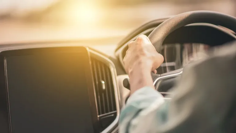 Driver's hand on the steering wheel of a right-hand-drive car, centre touchscreen to the left, low sun through the windscreen