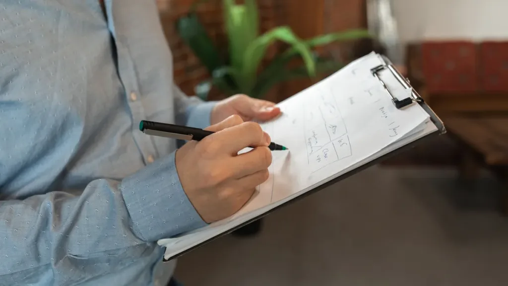 Person in a blue shirt writing notes with a pen on a clipboard, face out of frame, in an indoor room