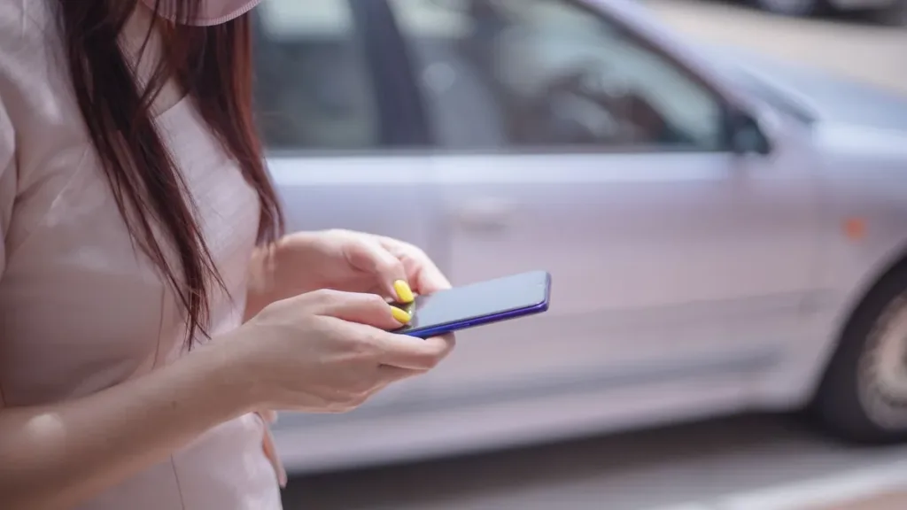 Close-up of a person's hands typing a message on a smartphone beside a parked silver car