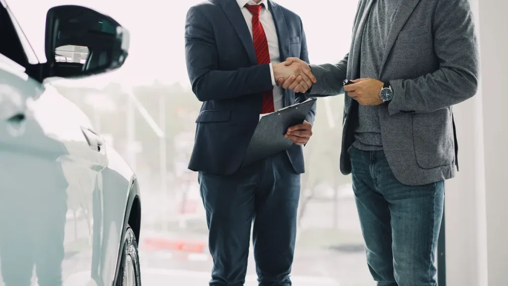 Two men in suits shaking hands beside a parked car, one holding a clipboard, faces out of frame