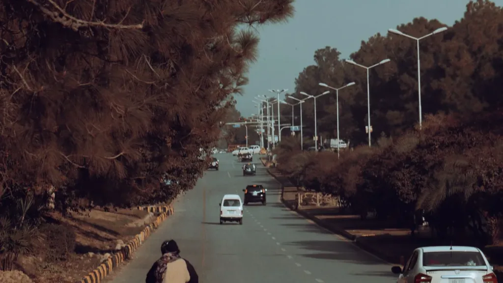 Tree-lined dual carriageway in Islamabad with a motorcyclist and a few cars travelling under autumn trees and street lamps