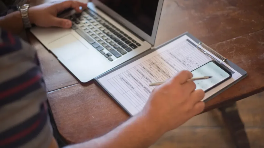 Hands filling in a form on a clipboard beside a smartphone and an open laptop on a wooden table