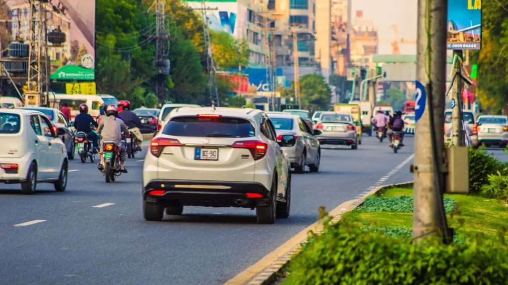 Cars and motorcycles driving along a tree-lined city road in Punjab, with a planted median and apartment blocks behind