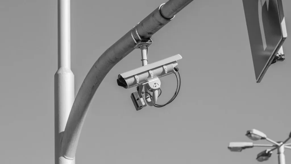 Black and white photo of a surveillance camera mounted on a curved street lamp pole beside a road sign