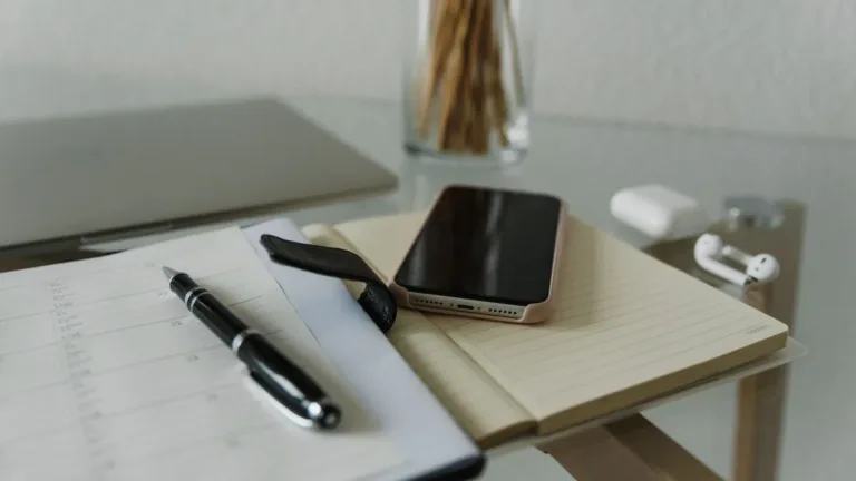 Smartphone resting on an open notebook beside a pen and wireless earphones on a glass desk