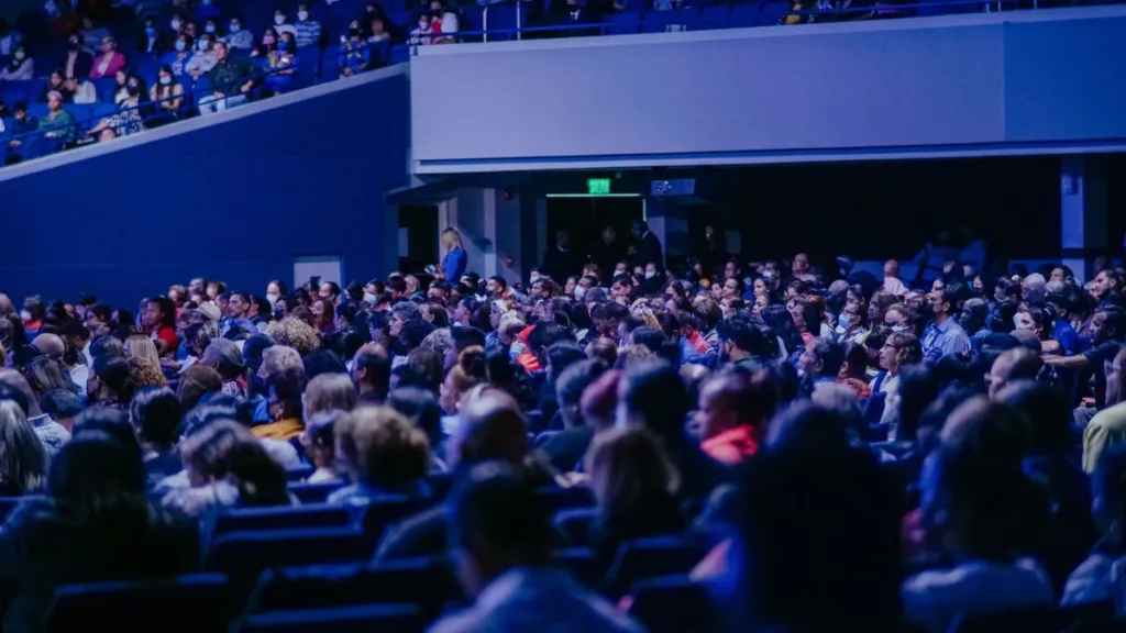 Rows of conference attendees seen from behind in a large auditorium with blue seats and an upper balcony