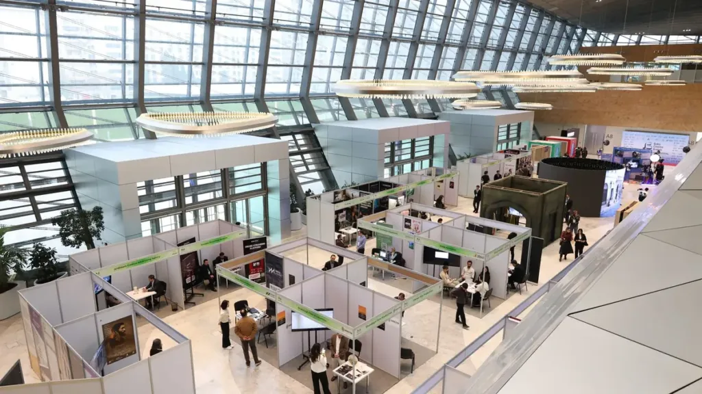 Elevated view of a glass-roofed exhibition hall with rows of white shell-scheme stands and visitors walking between them