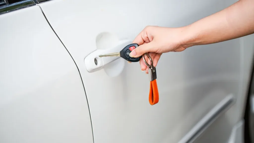 Close-up of a hand unlocking the door of a white car with a metal key and key fob on an orange strap
