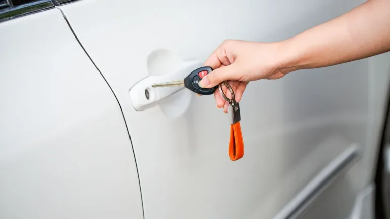 Close-up of a hand unlocking the door of a white car with a metal key and key fob on an orange strap