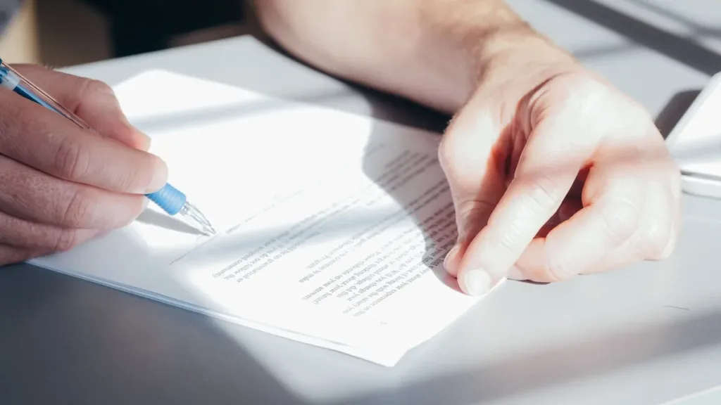 Close-up of hands signing a printed contract with a blue pen on a sunlit office desk