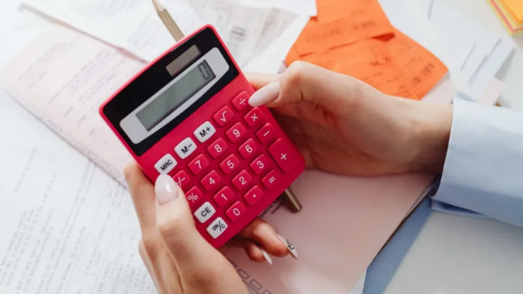 Hands holding a pink calculator over a desk covered in printed receipts and paper slips, tallying expenses