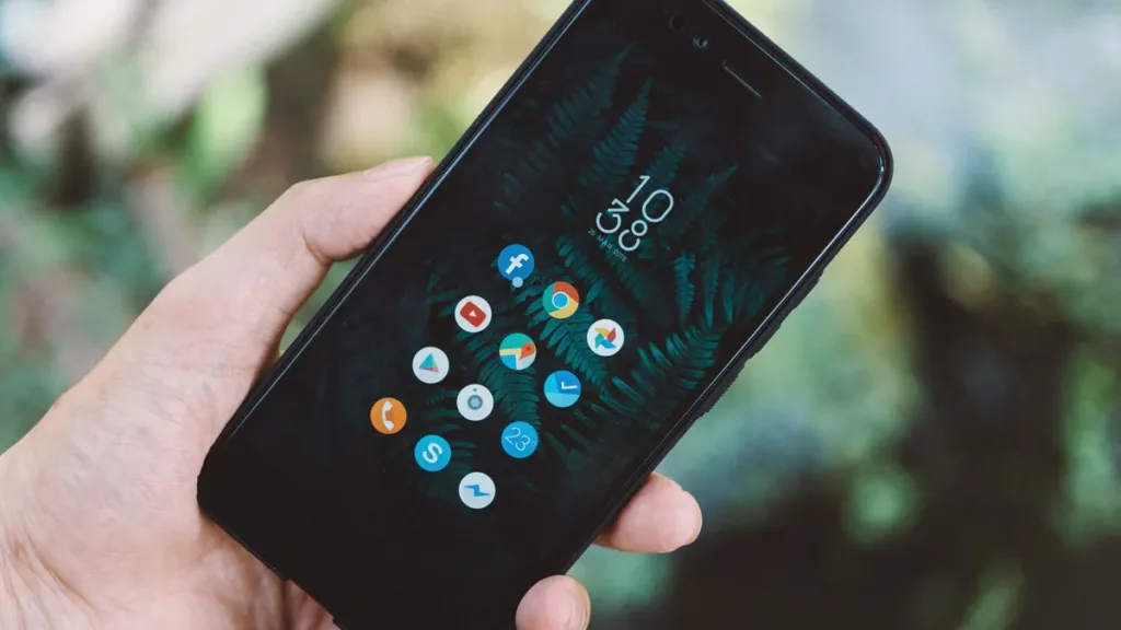 Hand holding a smartphone showing a home screen of app icons against a blurred green leafy background