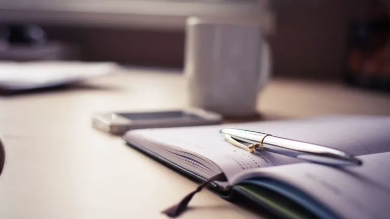 Open diary with a silver pen resting across its pages beside a phone and a white mug on a desk