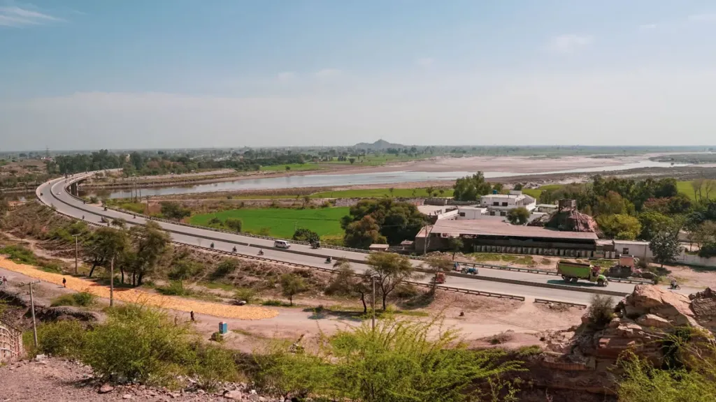 A divided highway curving through green countryside past low buildings and a wide river, seen from a hillside