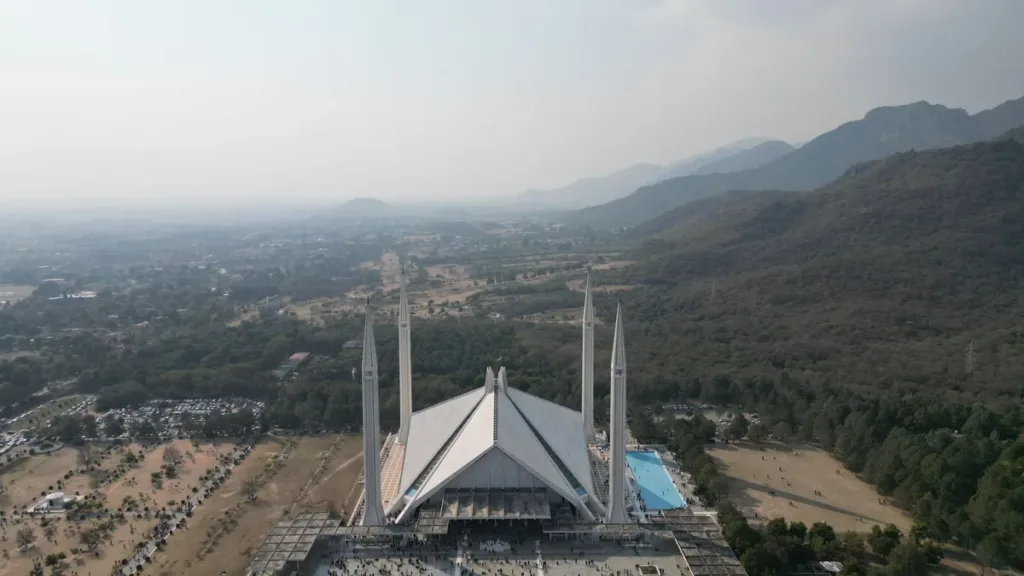Aerial view of Faisal Mosque's white minarets and tent-shaped prayer hall with the Margalla Hills behind, Islamabad