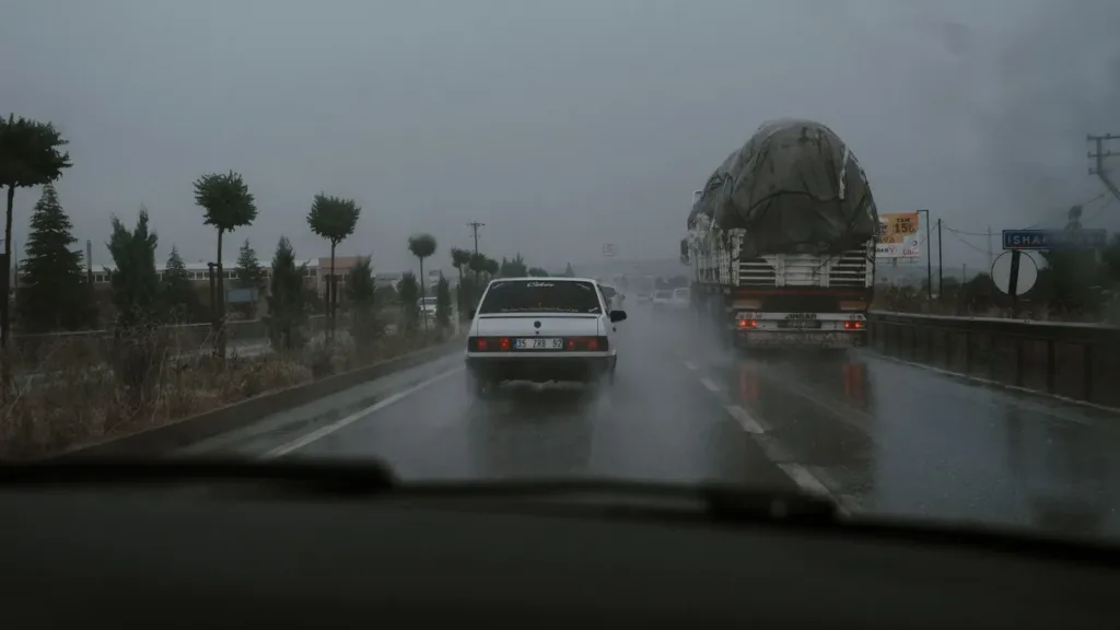 View through a rain-streaked windscreen of a car and a heavily loaded truck on a wet, grey highway